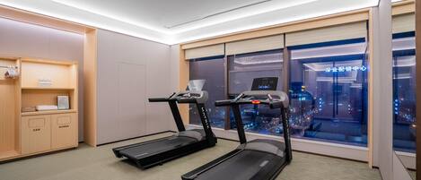 A modern hotel room with two treadmills, a wooden cabinet with toiletries, and a city view through large windows.