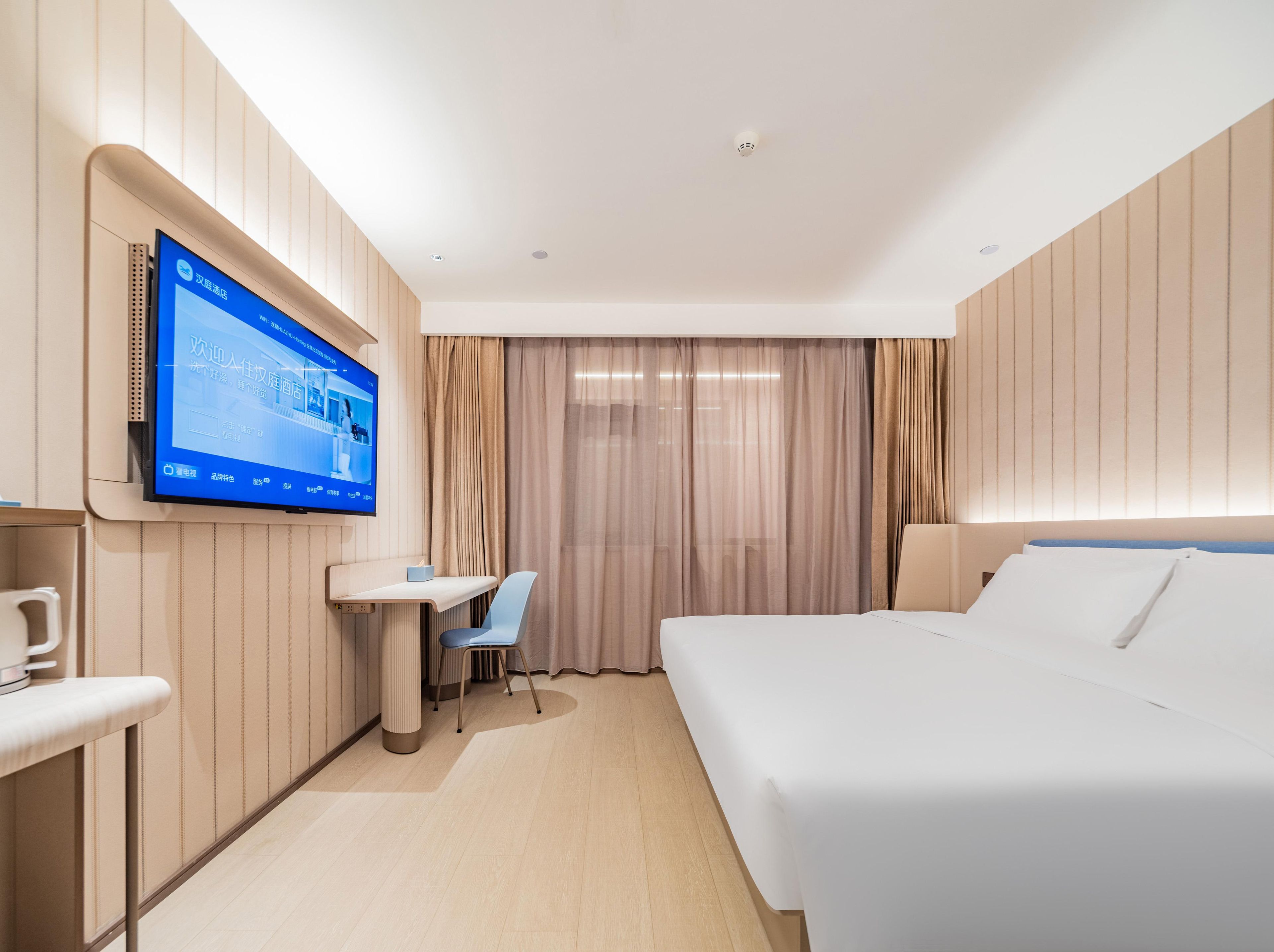 A hotel room with a bed, a desk, a chair, a television mounted on the wall, and a window with curtains.
