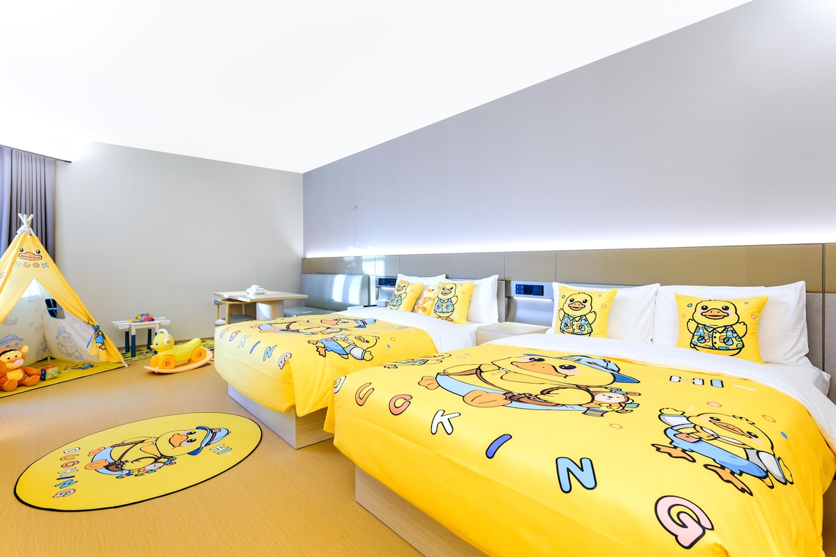 A hotel room with two beds, a desk, a chair, a small table, a yellow play tent, and a toy car.