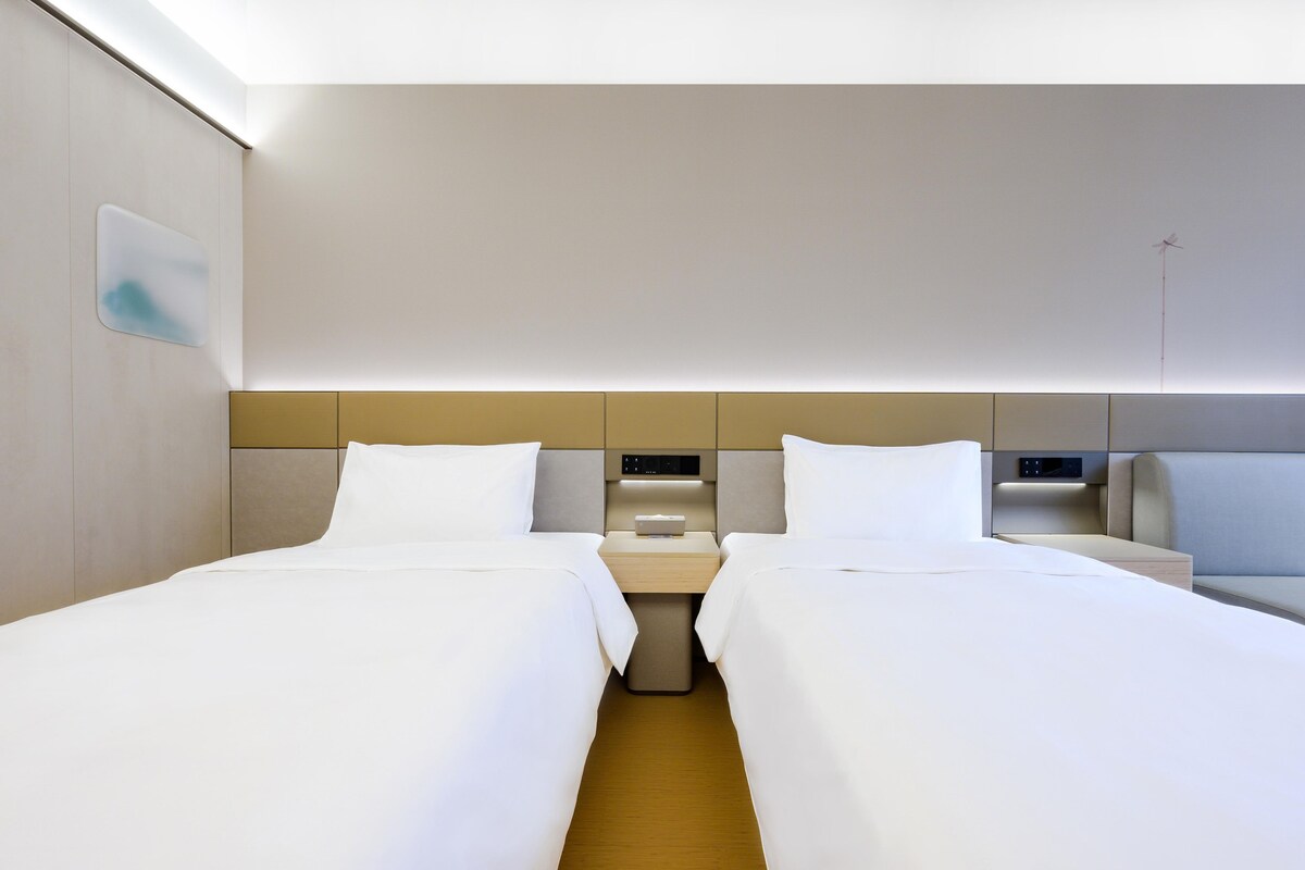 A hotel room with two single beds, a bedside table with a lamp, and a control panel on the wall.