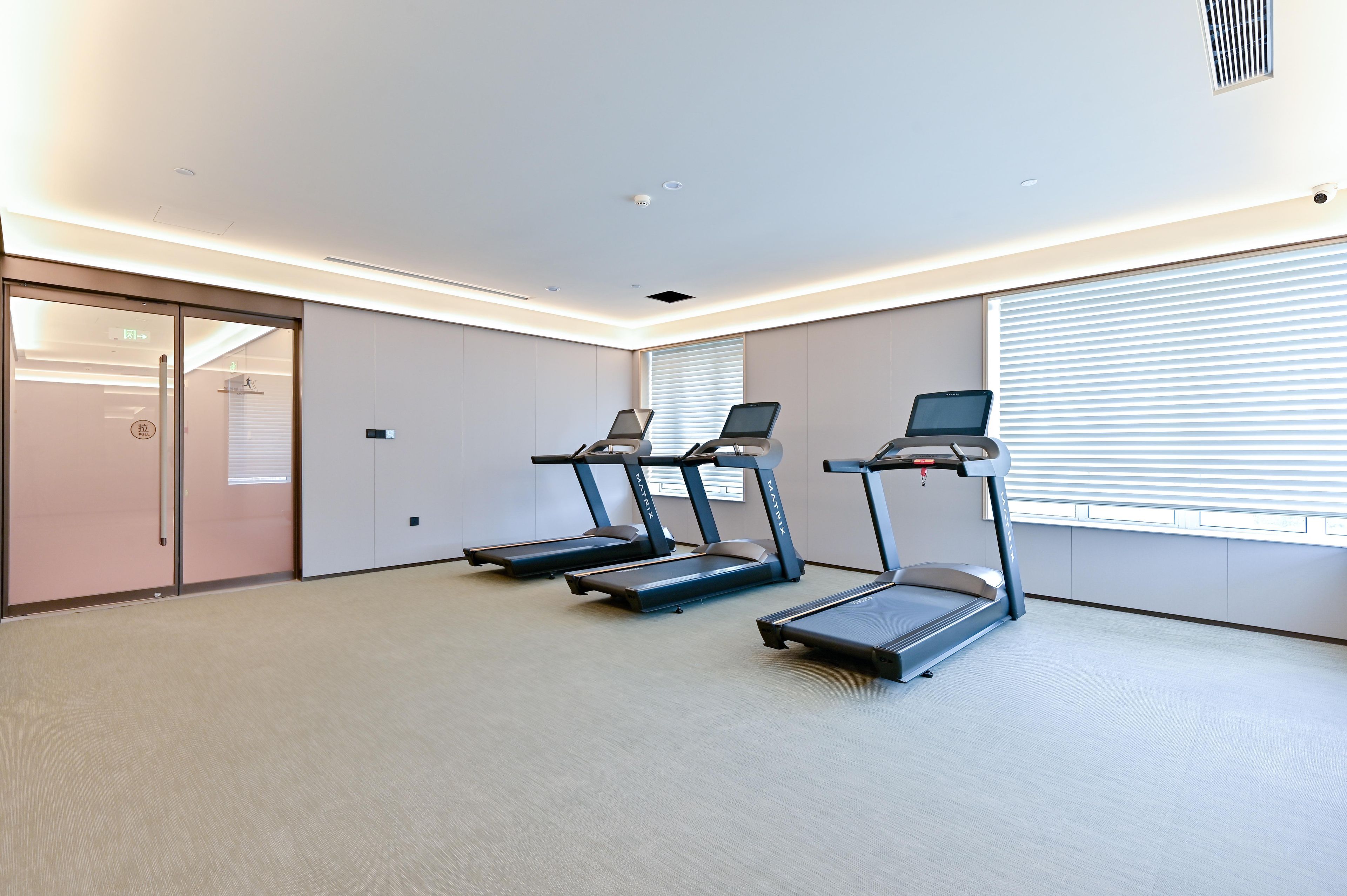 A fitness room with three treadmills, a glass door entrance, and a large window with blinds.