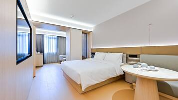 Habitación de hotel moderna con una cama grande, una mesita de noche con tetera, una zona de estar con sofá y un ventanal con cortinas.