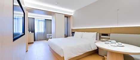 Habitación de hotel moderna con una cama grande, una mesita de noche con tetera, una zona de estar con sofá y un ventanal con cortinas.