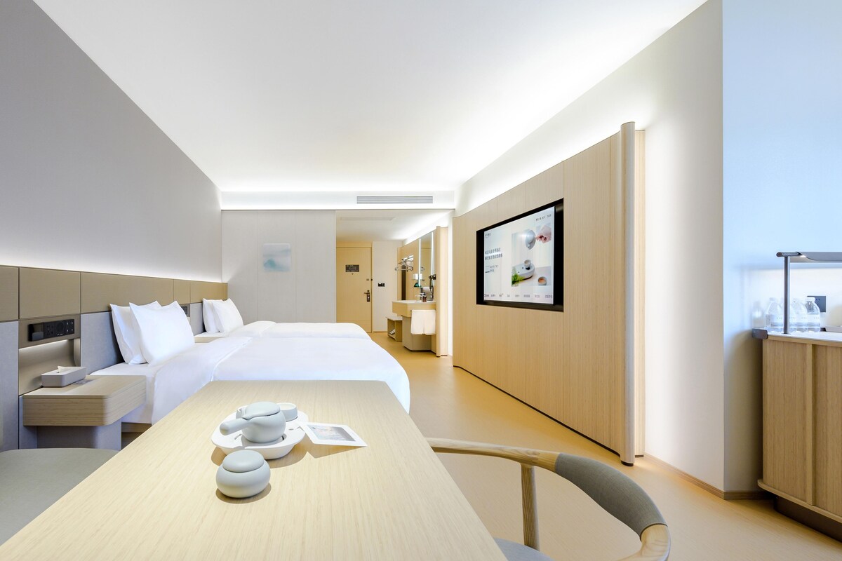 A modern hotel room with a large bed, bedside tables, a desk with a chair, a bathroom with a mirror, and a wall-mounted TV.