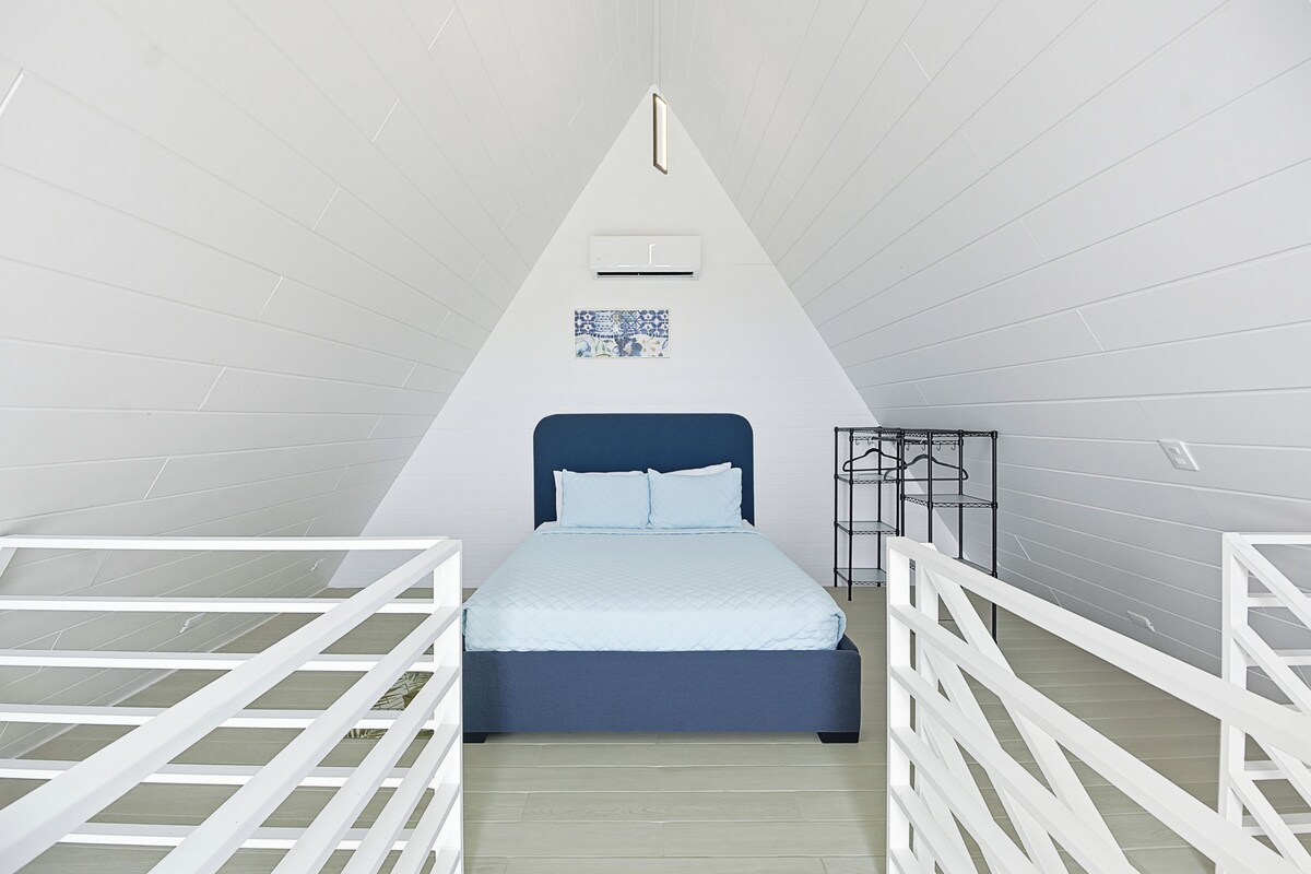 Habitación triangular con cama centrada, barandales blancos, una pequeña estantería y aire acondicionado instalado en el techo.