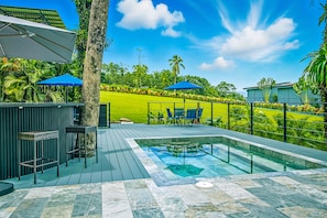 A pool area with a hot tub, seating, and a view of a well-maintained lawn and trees.