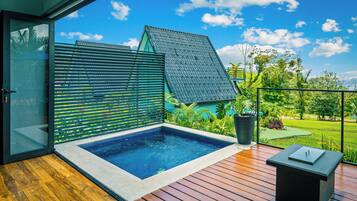 A modern outdoor swimming pool with a wooden deck, a glass door leading to a covered area, and a view of a lush garden and a building with a sloped roof.