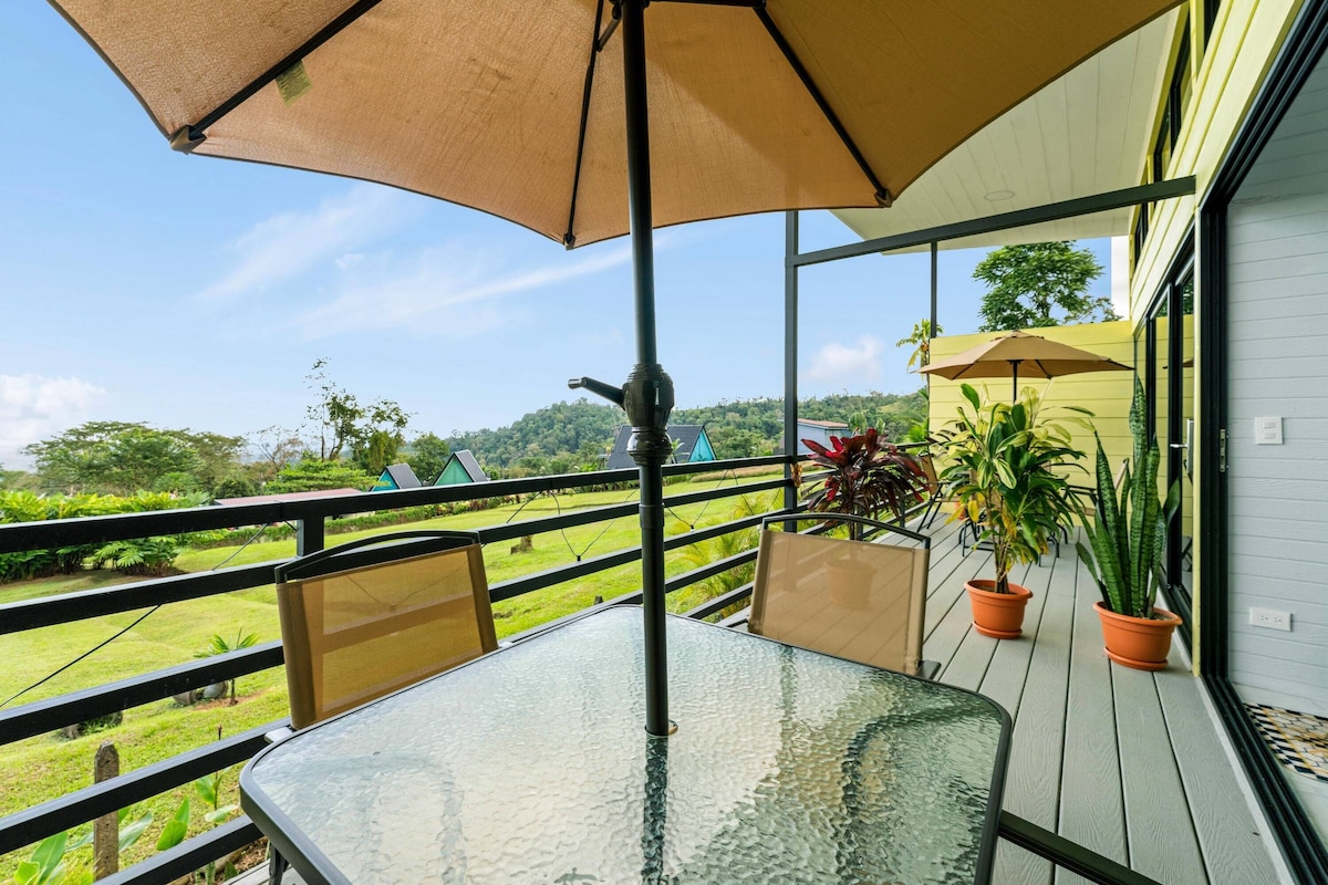 A glass table with a beige umbrella, outdoor chairs, potted plants, and a view of greenery and hills.