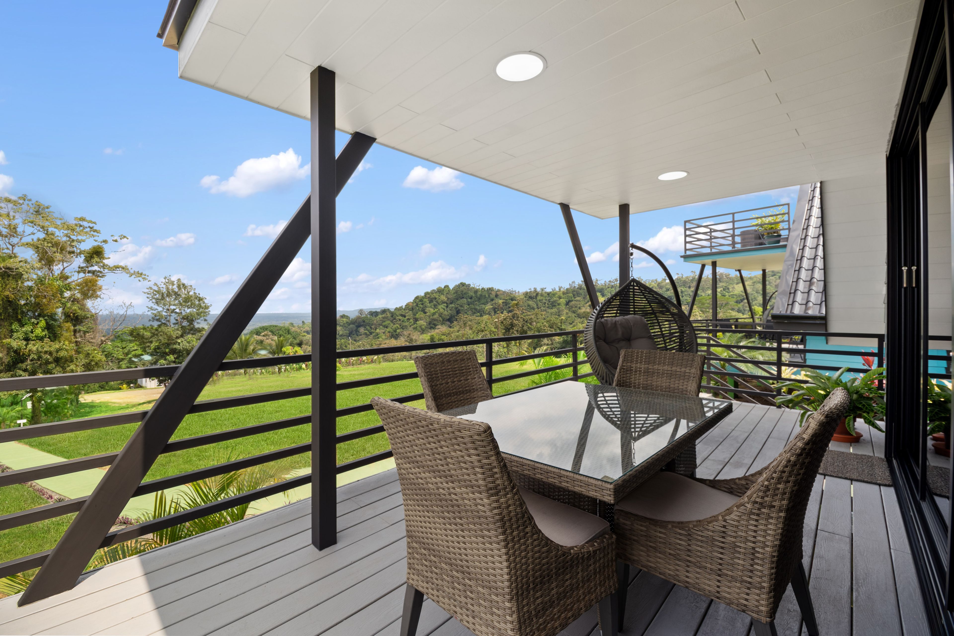 A balcony with a glass table and wicker chairs, overlooking a green landscape.