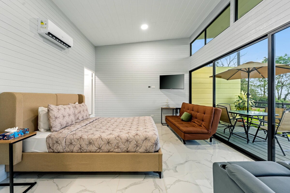 Un dormitorio moderno con cama, sofá, televisor y balcón con mobiliario exterior.