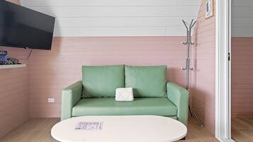 Une pièce avec un canapé vert, une table blanche, un téléviseur fixé au mur et un porte-manteau.