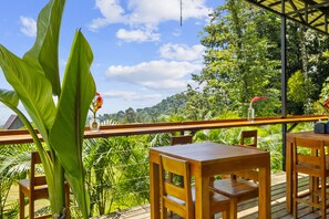 Un espacio exterior con mesas y sillas de madera, rodeado de vegetación exuberante y vistas a una colina.
