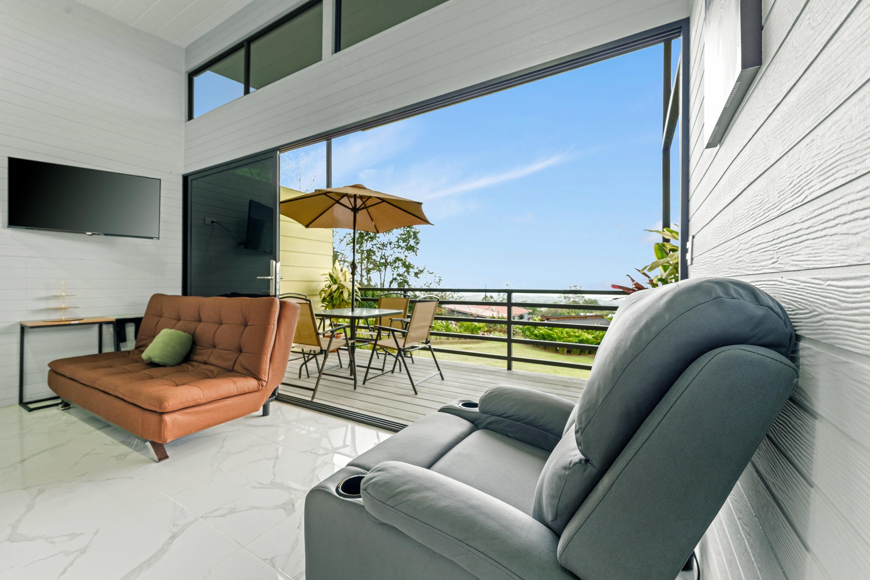 Un salon moderne avec un canapé, un téléviseur fixé au mur et un balcon agrémenté de mobilier d’extérieur.