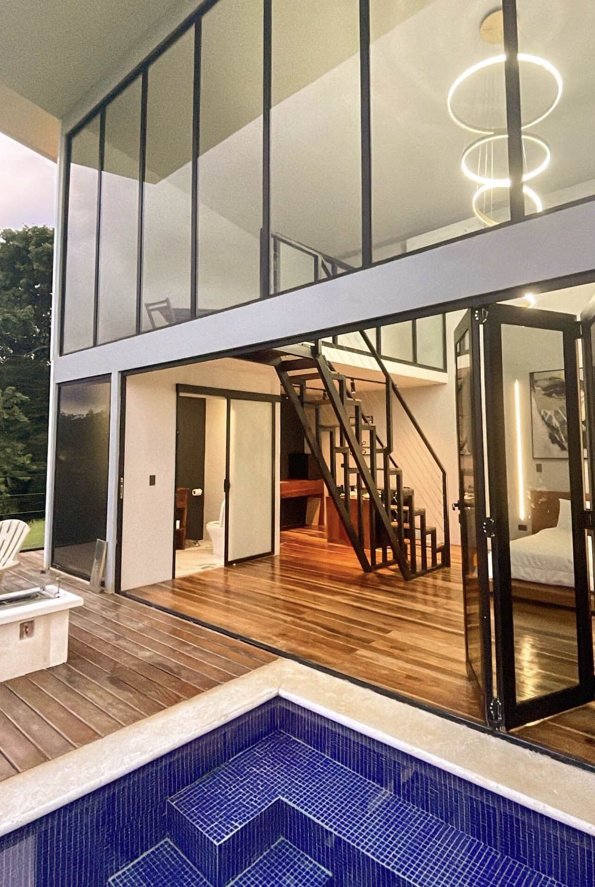 A modern two-story house with a visible staircase leading to the upper floor, a pool area with blue tiles, and a wooden deck.