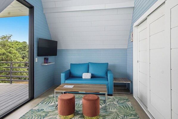 A balcony with a blue sofa, a wooden table, two stools, and a mounted TV.
