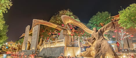 A night scene with a water fountain, stone statues, and traditional architecture.