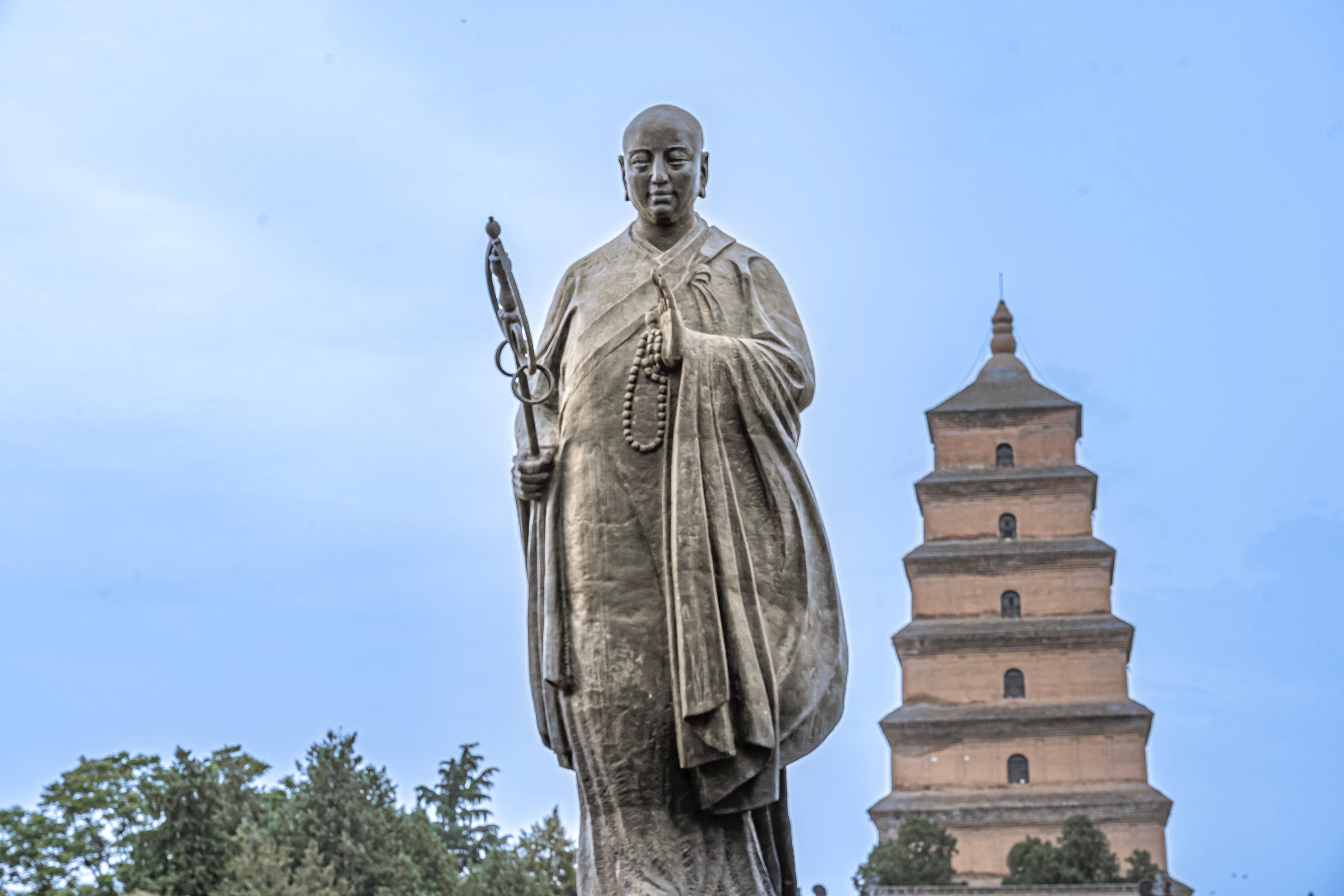 Una estatua imponente de una figura histórica sosteniendo un báculo y un rosario, con una torre de estilo pagoda al fondo.