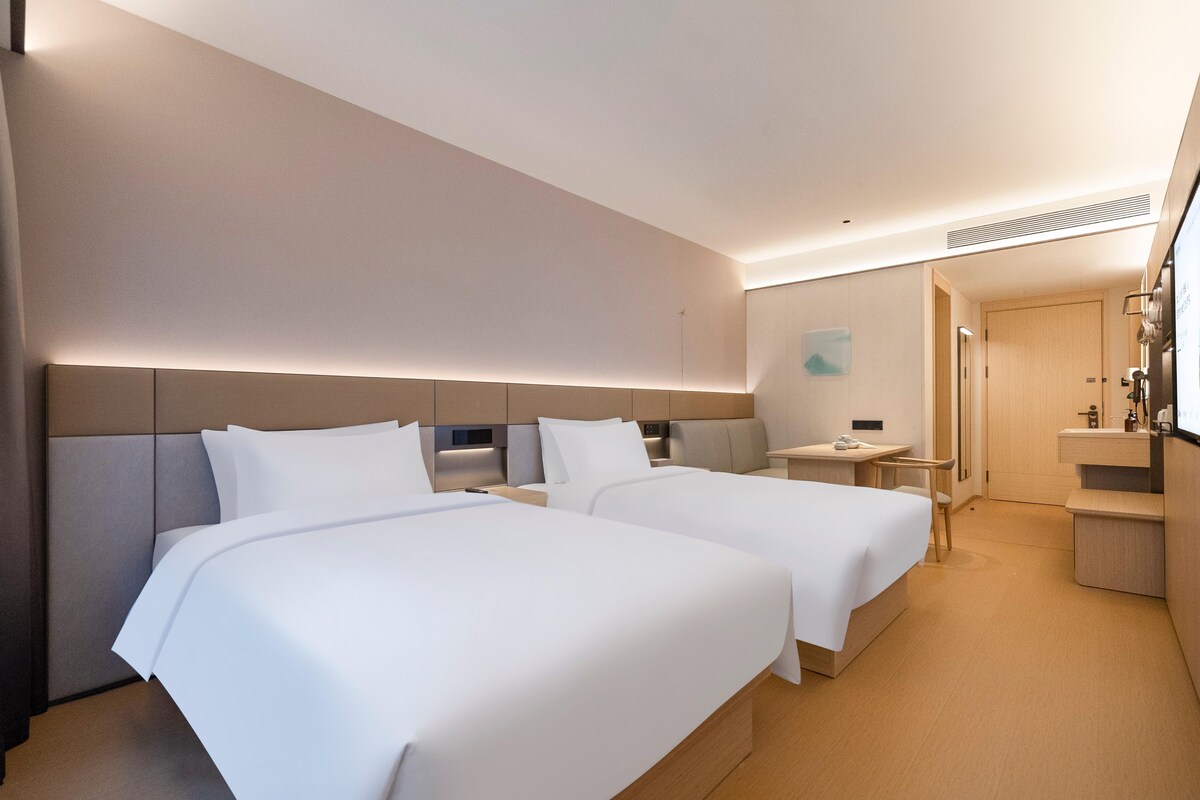 A hotel room with two beds, a small table, a chair, a desk, and a bathroom.