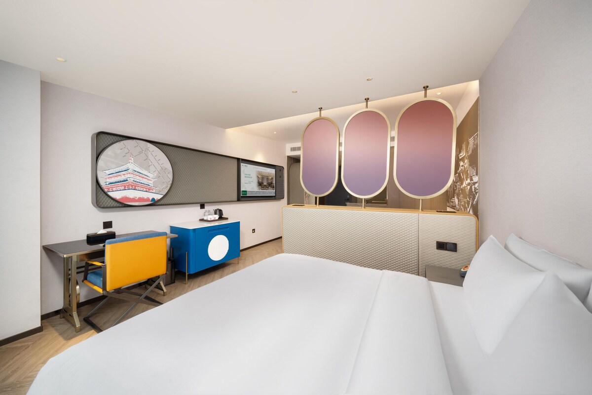 A modern hotel room with a large bed, a desk, a chair, a vanity, and a wall-mounted TV.