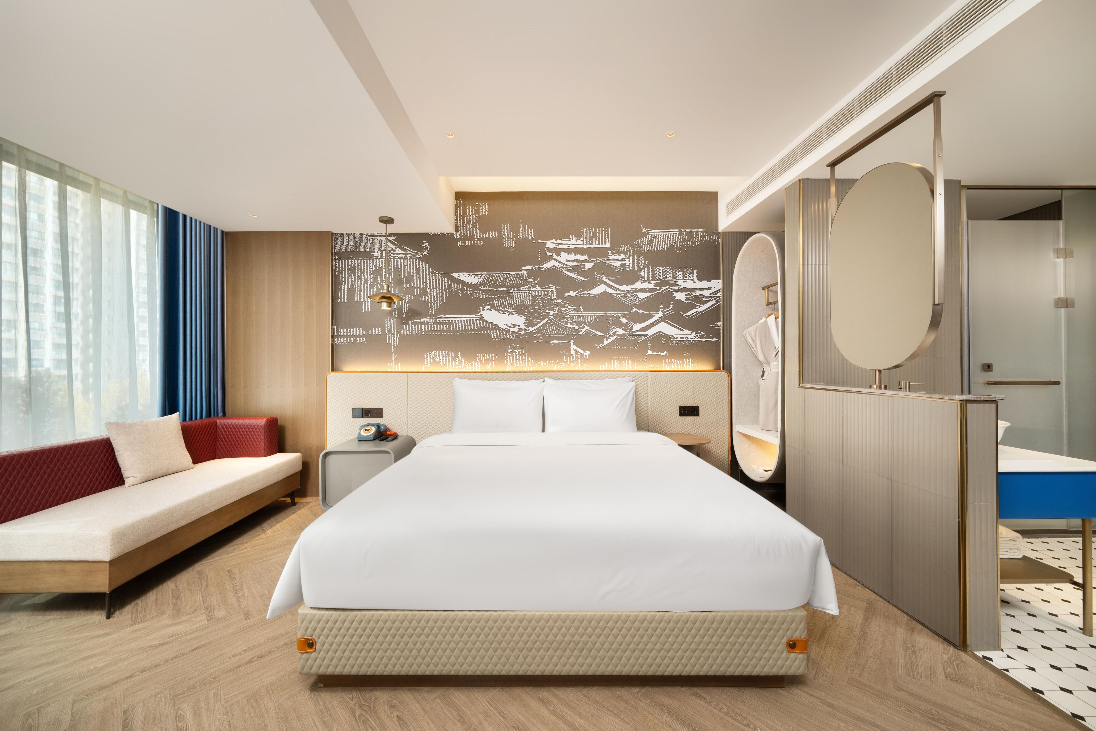 A hotel room with a large bed, a sofa, a desk, a mirror, and a wall with a cityscape mural.