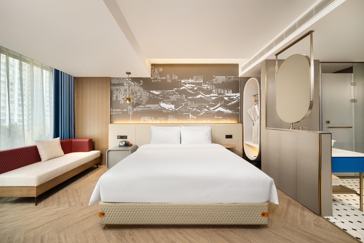 A hotel room with a large bed, a sofa, a desk, a mirror, and a wall with a cityscape mural.