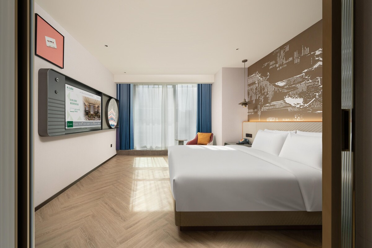 A hotel room with a bed, a desk with a lamp, a chair, a large window with curtains, and a wall-mounted TV.