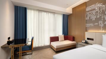 A hotel room with a bed, a desk, a chair, a sofa, and a large window with curtains.