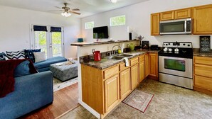 A compact kitchen with wooden cabinets, a microwave, a stove, and a dining area with a blue sofa and a red throw blanket. - Pura Vida Biloxi (Biloxi)