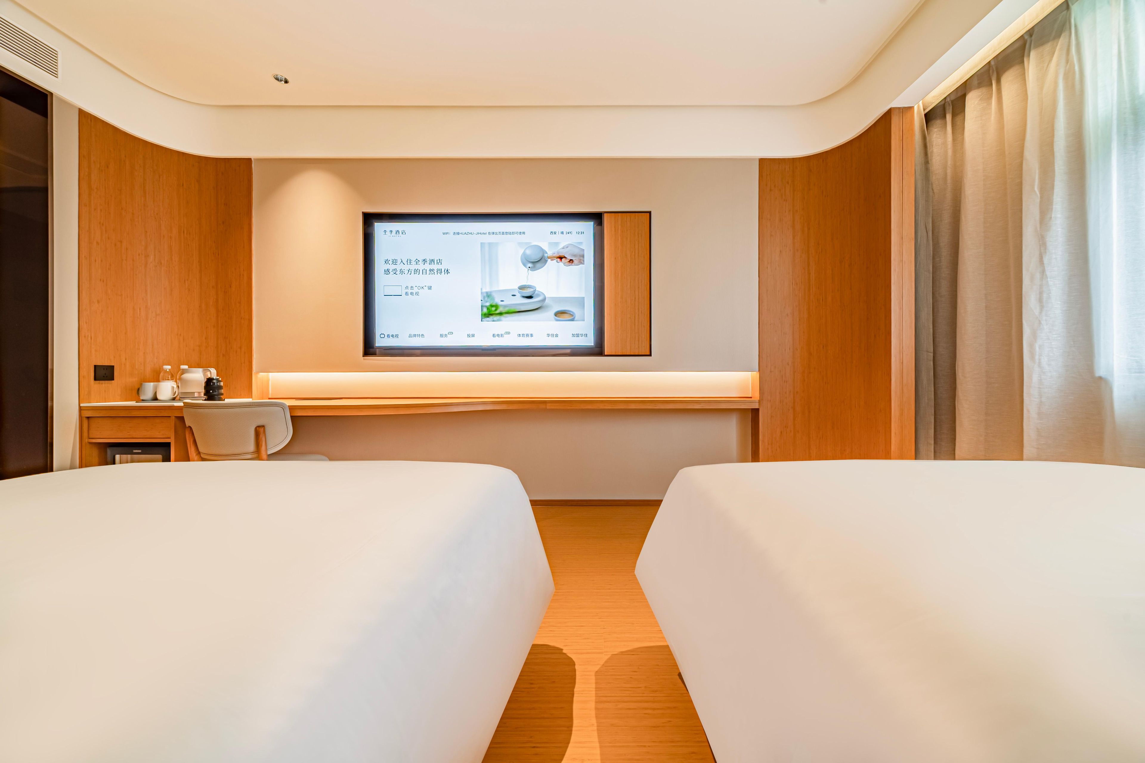 A hotel room with two beds, a desk with a computer, and a large screen displaying a hotel website.