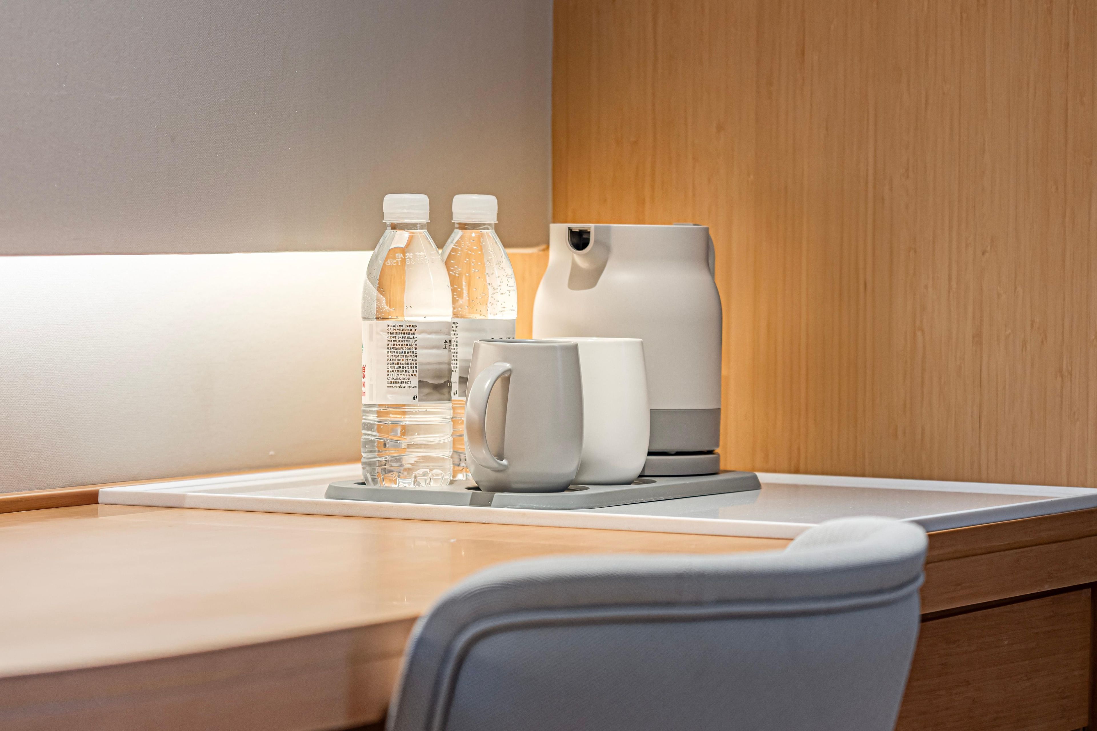 A clean, modern hotel room with a wooden desk, a white mug, two water bottles, and a coffee maker.