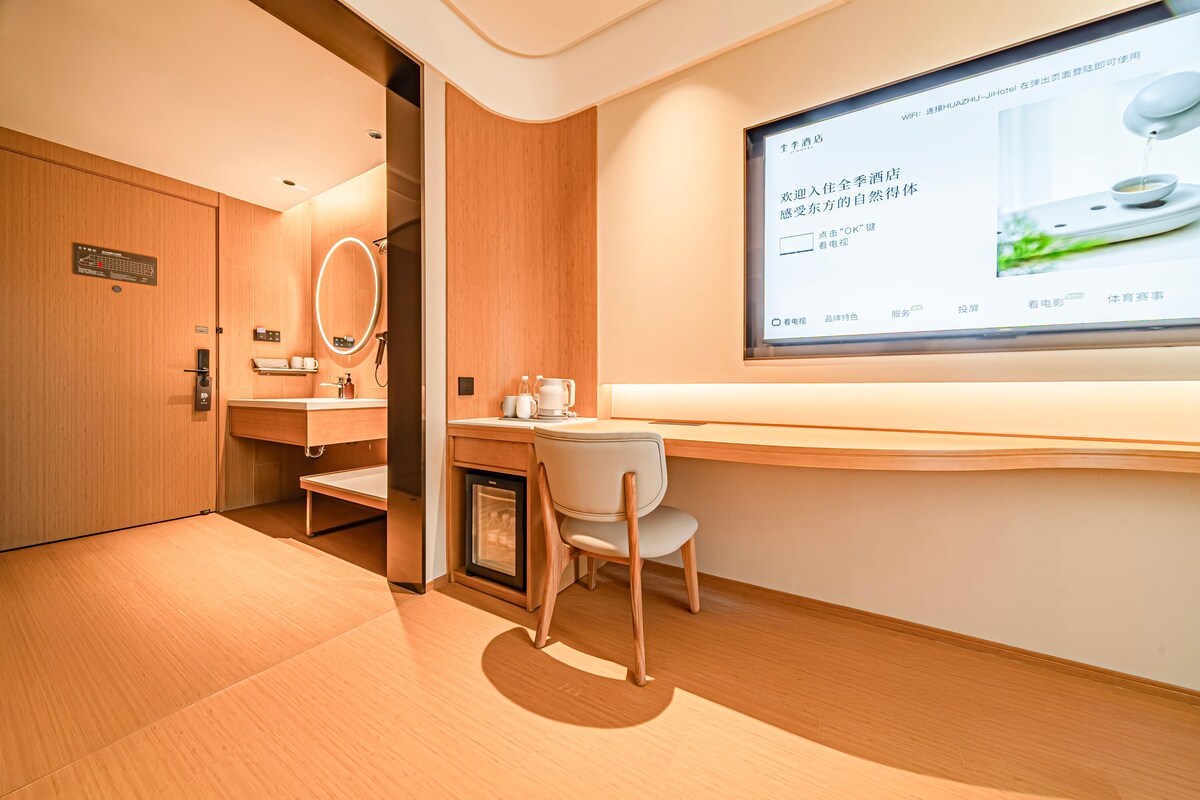 A modern hotel room with a wooden floor, a desk with a chair, a small fridge, a vanity with a mirror, and a large screen displaying information.