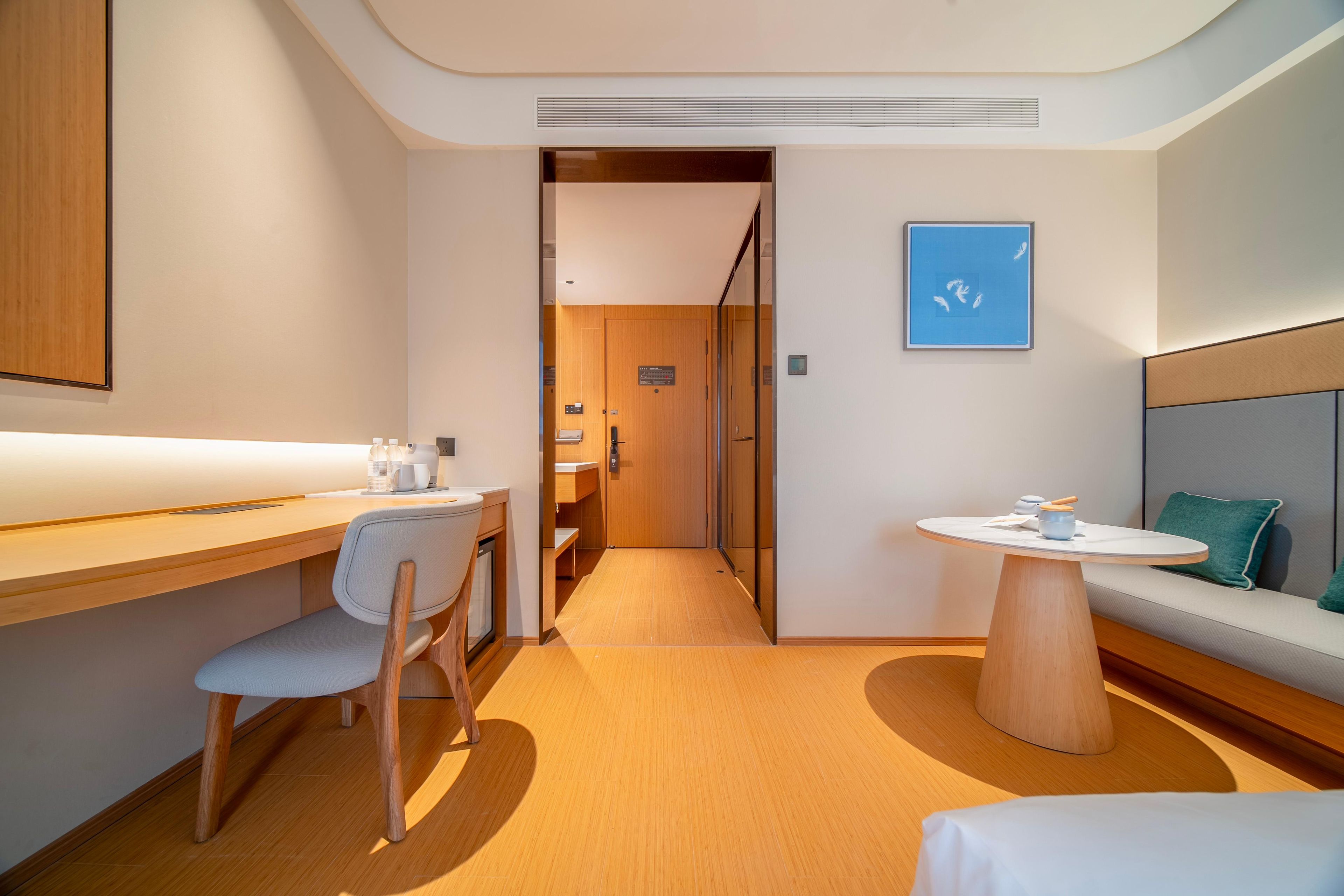 A modern hotel room with a wooden desk, a chair, a round table, and a sofa with a cushion.