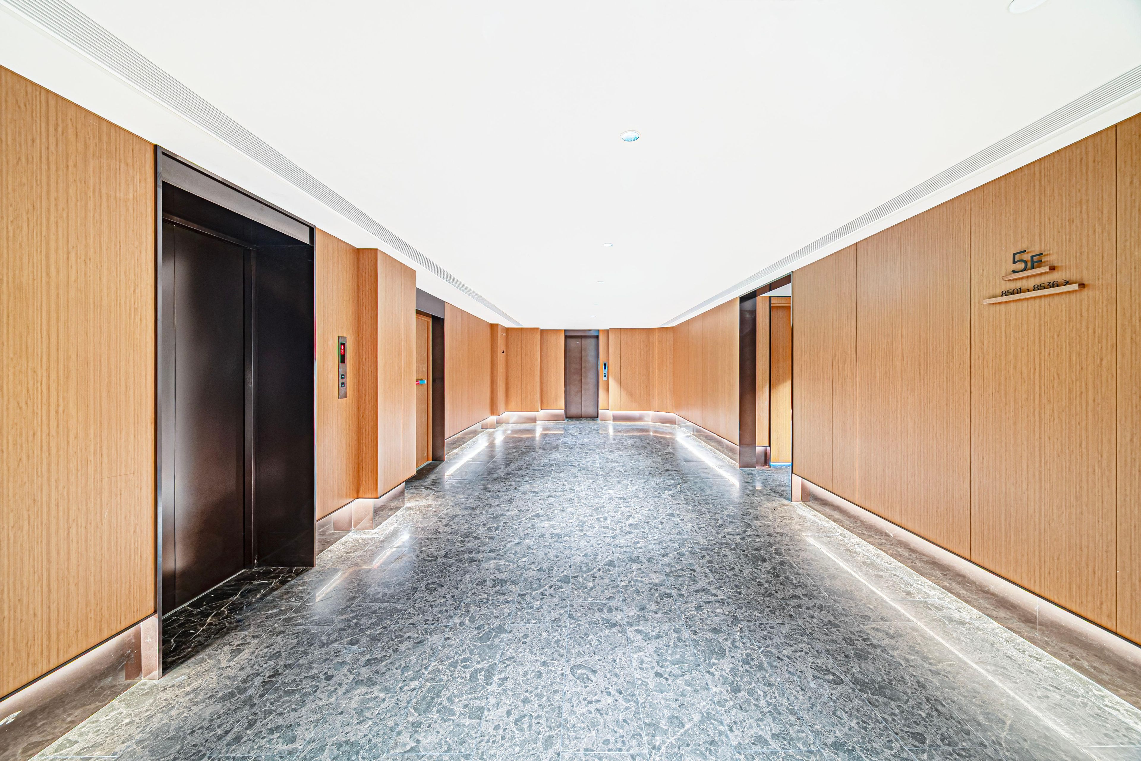 A modern hallway with wooden paneling and a marble floor, leading to a door with a digital elevator control panel.