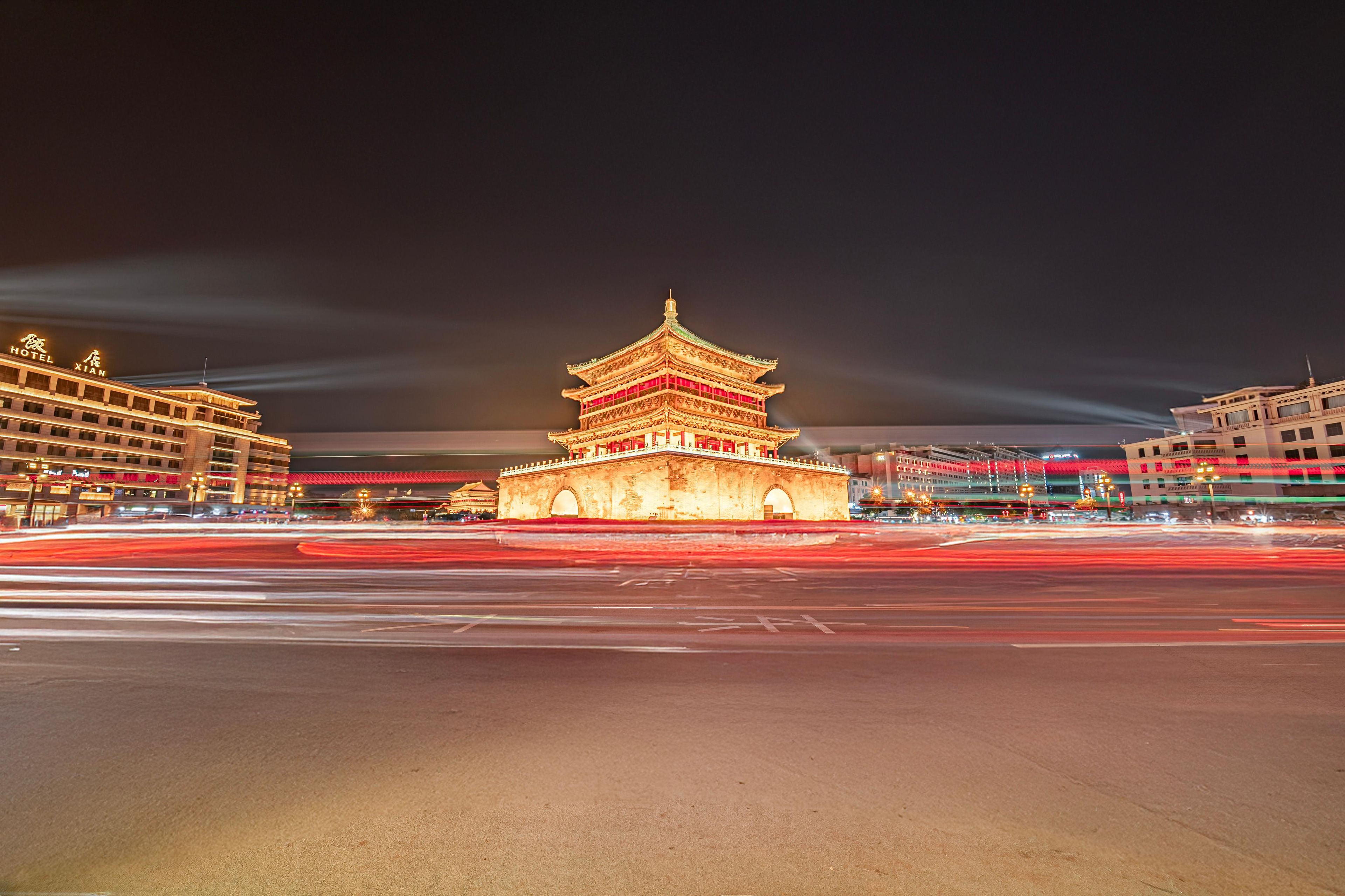 A traditional Chinese building with multiple tiers and a red roof, surrounded by modern buildings with illuminated signage.