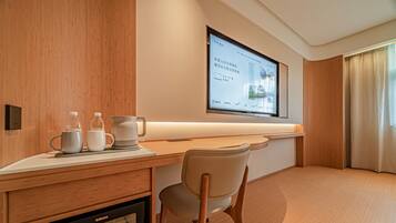 A hotel room with a wooden desk, a chair, a television mounted on the wall, and a window with curtains.