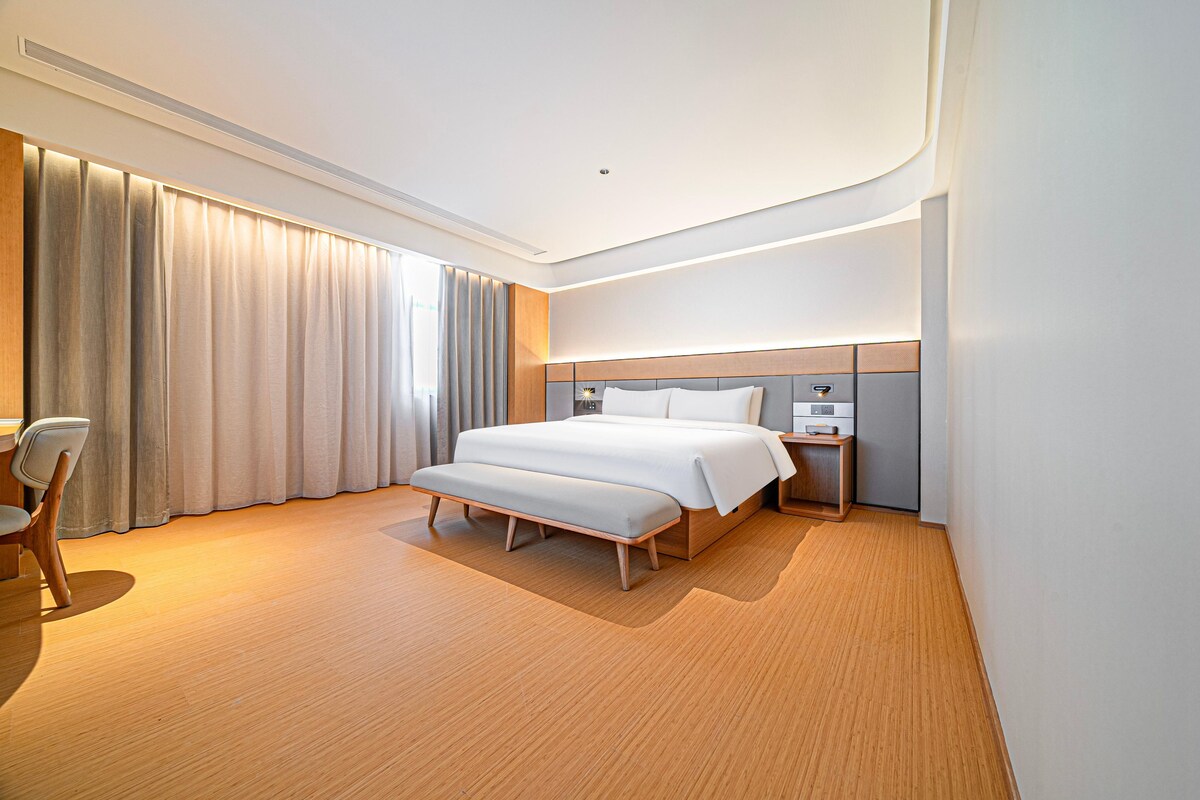 Habitación de hotel moderna con una cama grande, una mesita de noche, una silla y un amplio ventanal con cortinas.