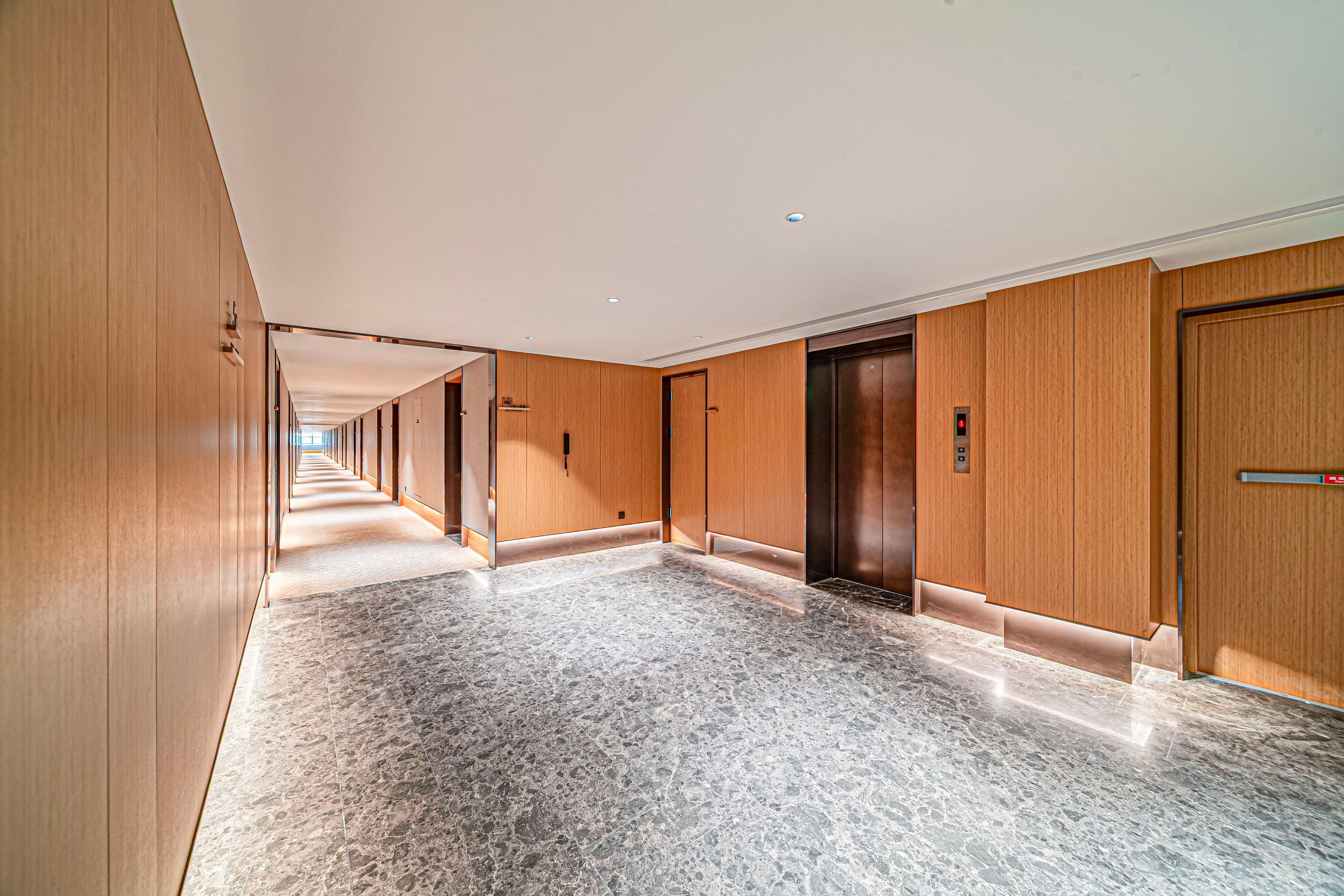 A long corridor with wooden paneling and marble flooring, leading to an elevator and a door.