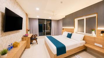 A modern hotel room with a flat-screen TV, a desk with a chair, a bed with a blue blanket, and a large window.