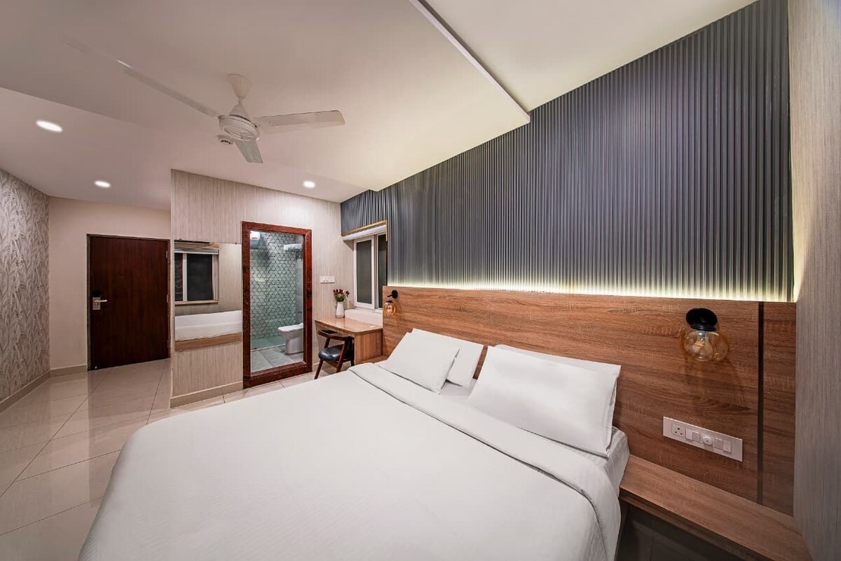 A modern hotel room with a large bed, a desk, a vanity, and a shower area.