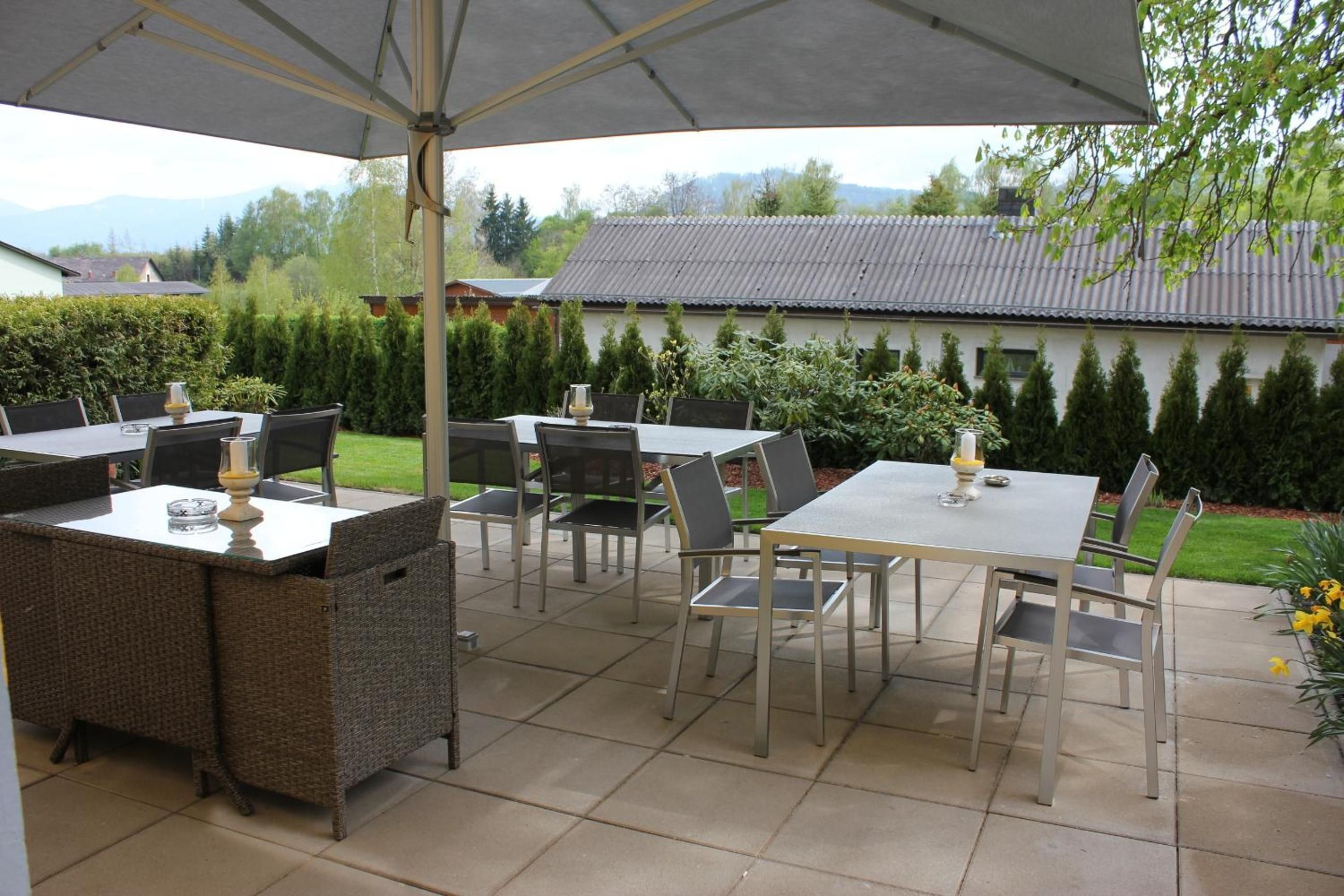 Un patio al aire libre con mesas y sillas, una zona cubierta y vista a una casa y árboles de fondo.