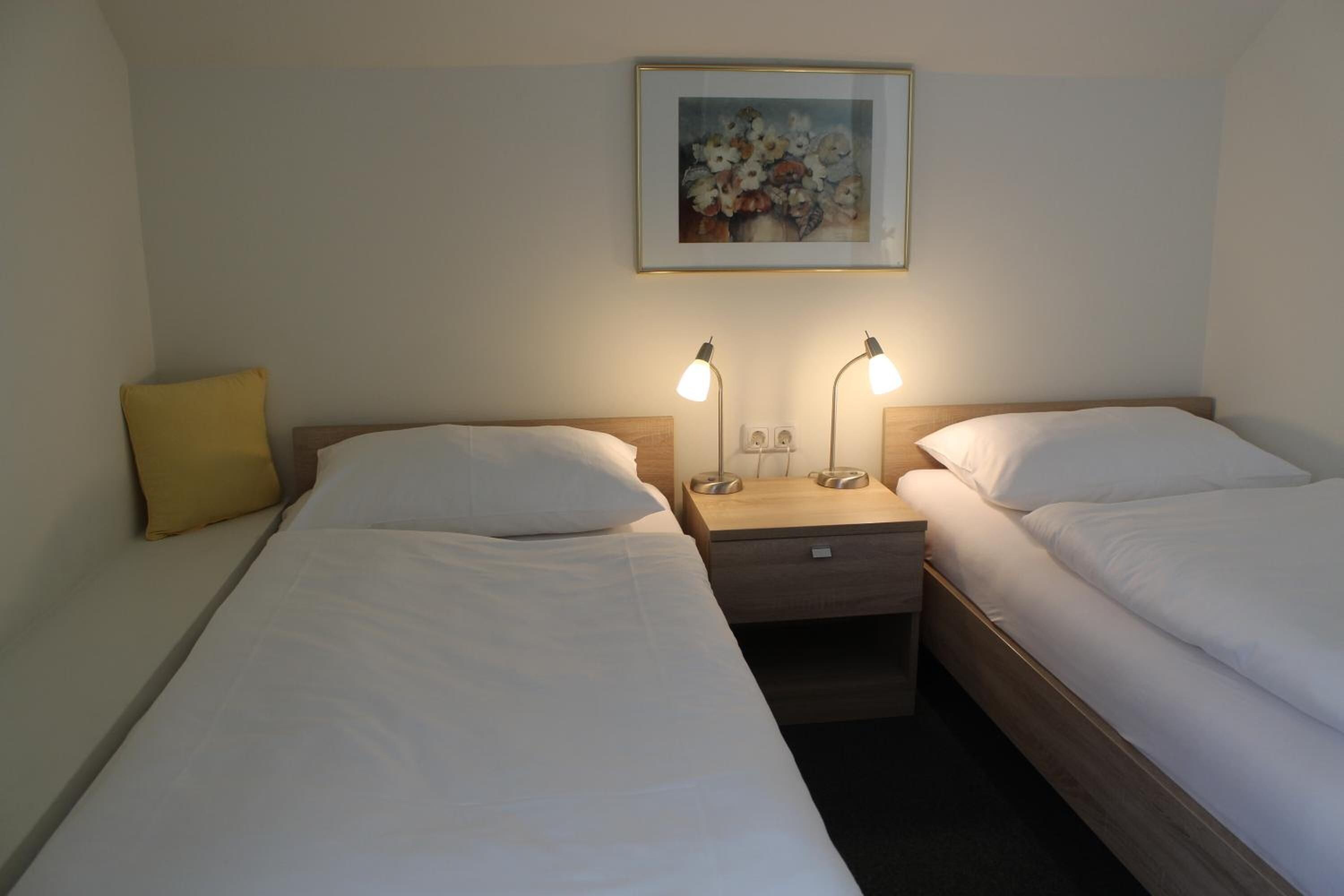 A hotel room with two single beds, a nightstand with lamps, a painting on the wall, and a yellow pillow.