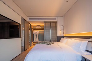 Ein Hotelzimmer mit einem großen Bett, einem Nachttisch, einem Fernseher, einem Kleiderschrank und einem durch eine Glastür einsehbaren Badezimmer.