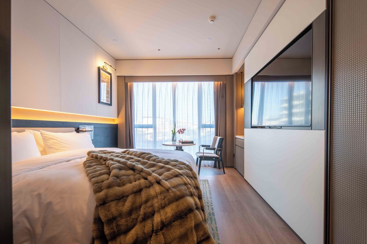 A hotel room with a large bed, a view of the city, and a dining area with a table and chairs.
