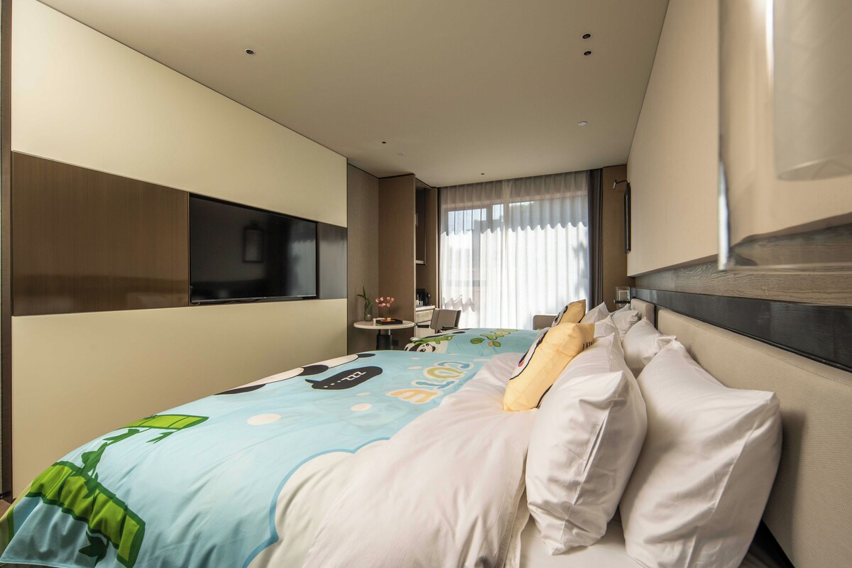 A hotel room with a bed, a television, a small table with a vase, and a window with curtains.