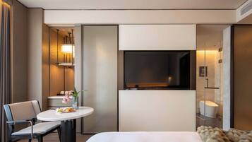Habitación de hotel con una cama, una mesa redonda con sillas, un televisor montado en la pared y un baño visible a través de una puerta corredera.