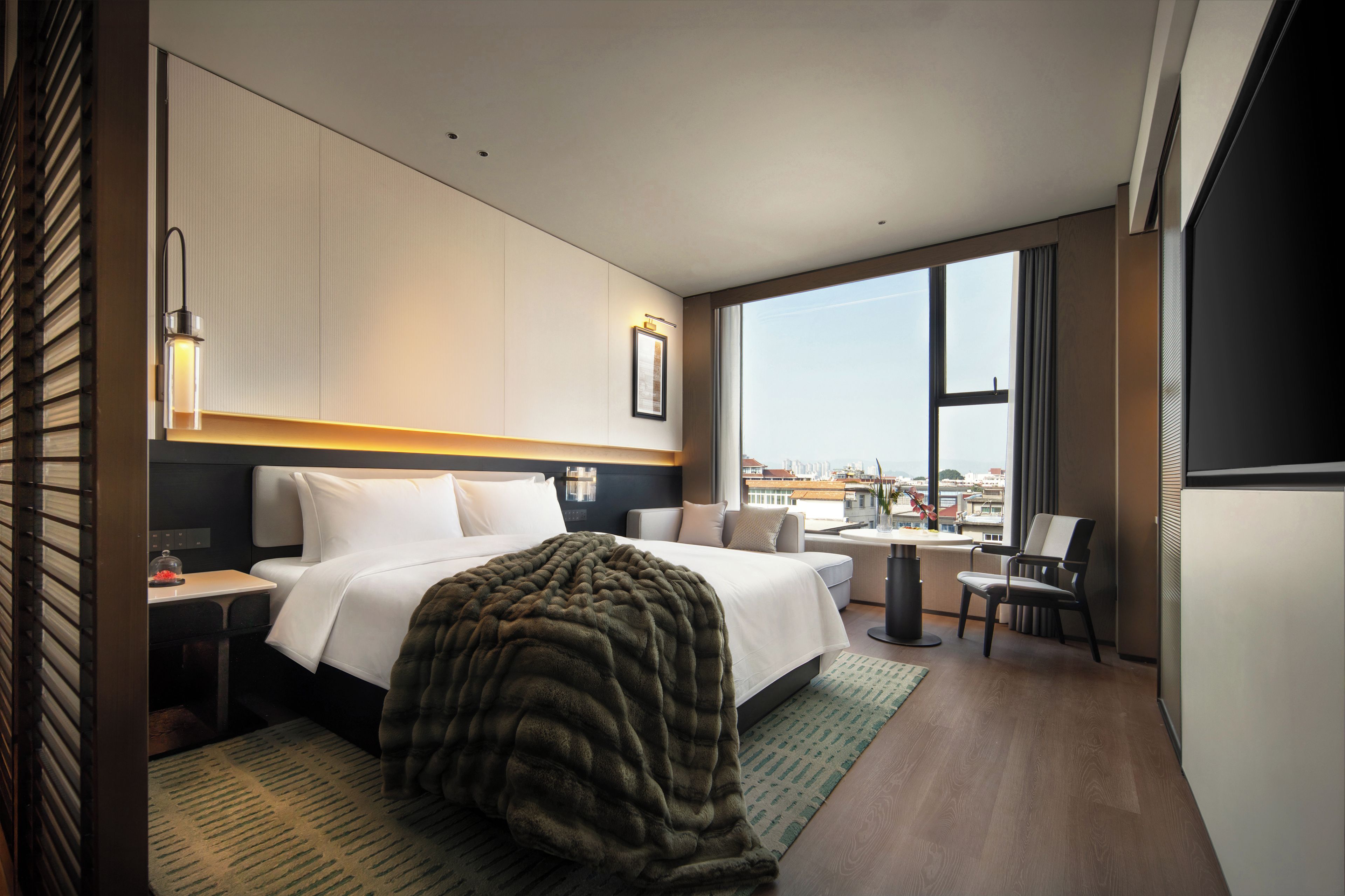 Habitación de hotel moderna con una cama grande, un escritorio, una silla y vistas de la ciudad a través de ventanas amplias.