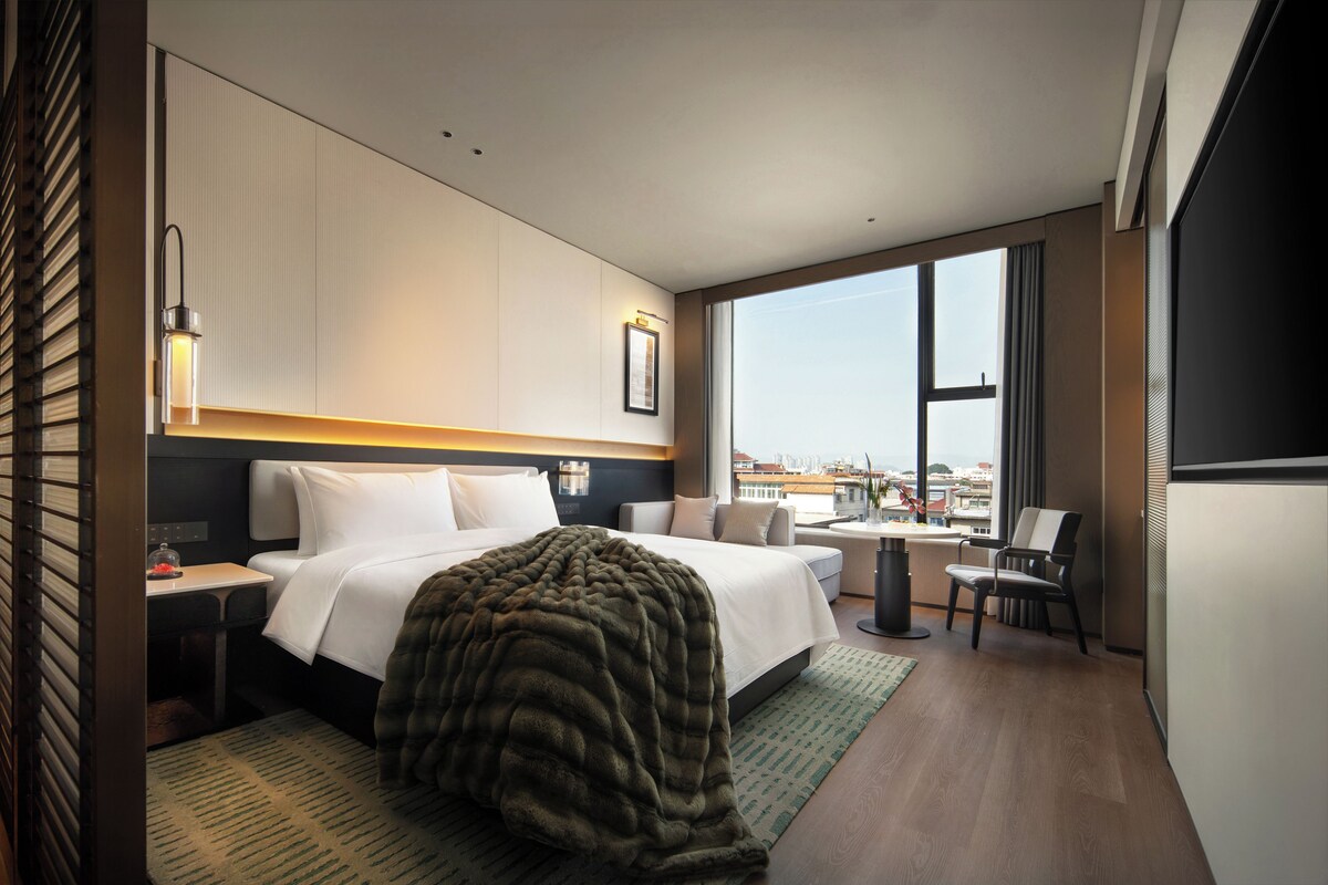 A modern hotel room with a large bed, a desk, a chair, and a view of the city through large windows.