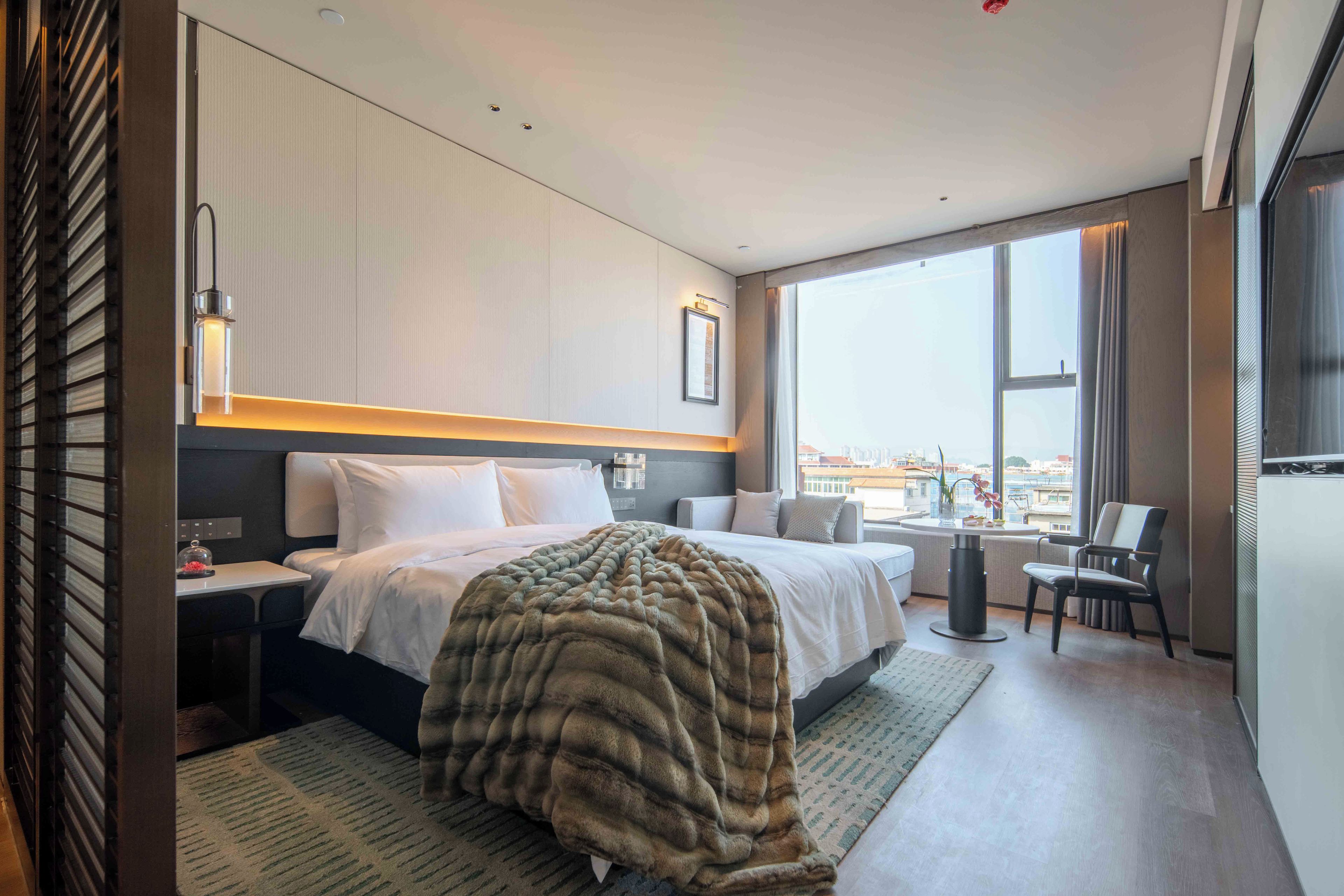 Habitación de hotel moderna con una cama grande, un escritorio, una silla y vistas de la ciudad a través de amplios ventanales.