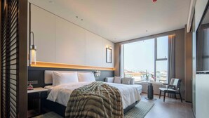 A modern hotel room with a large bed, a desk, a chair, and a view of the city through large windows.