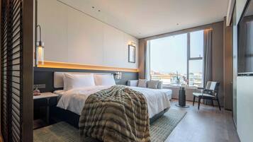 Habitación de hotel moderna con una cama grande, un escritorio, una silla y vistas de la ciudad a través de amplios ventanales.